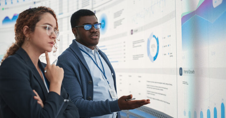 Two colleagues talking and gesturing in front of a big screen showing business analytics and AI tools.